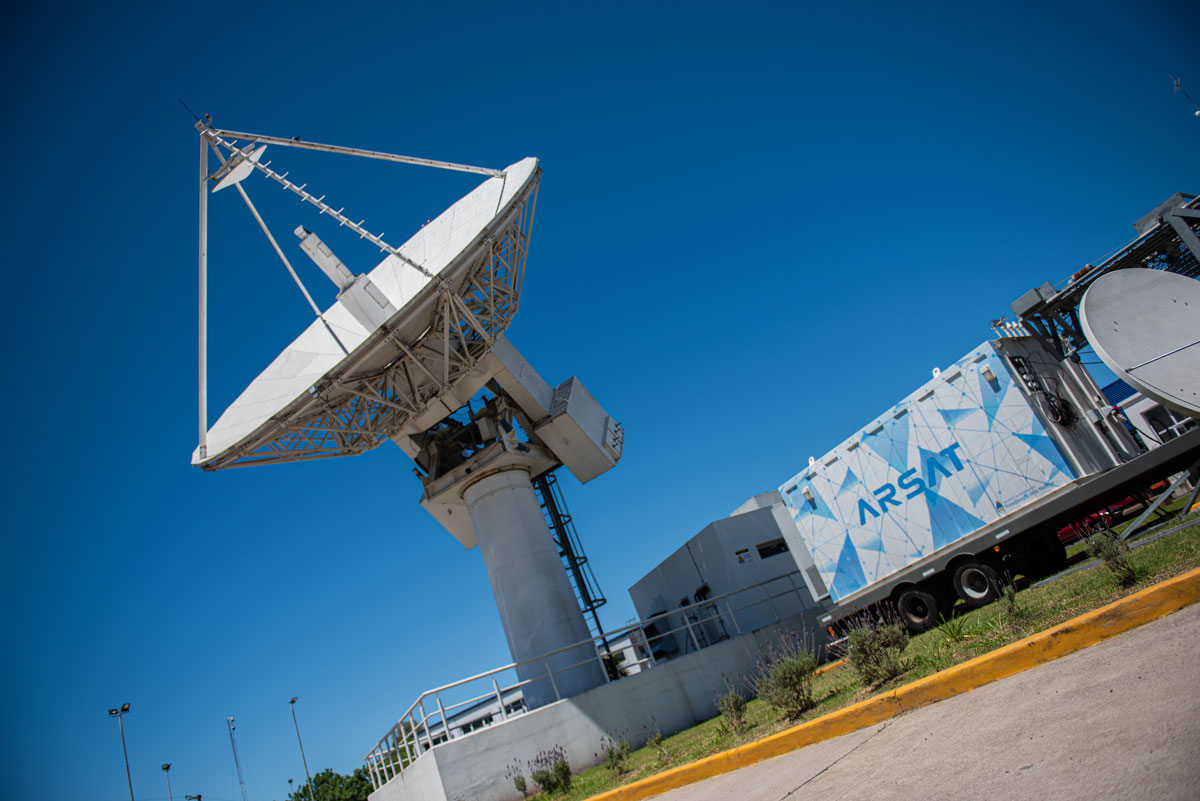 Una gran antena de la Estación Terrena de Benavídez, que apunta hacia el cielo donde están el Arsat 1 y 2, junto a un shelter con el logo de Arsat.